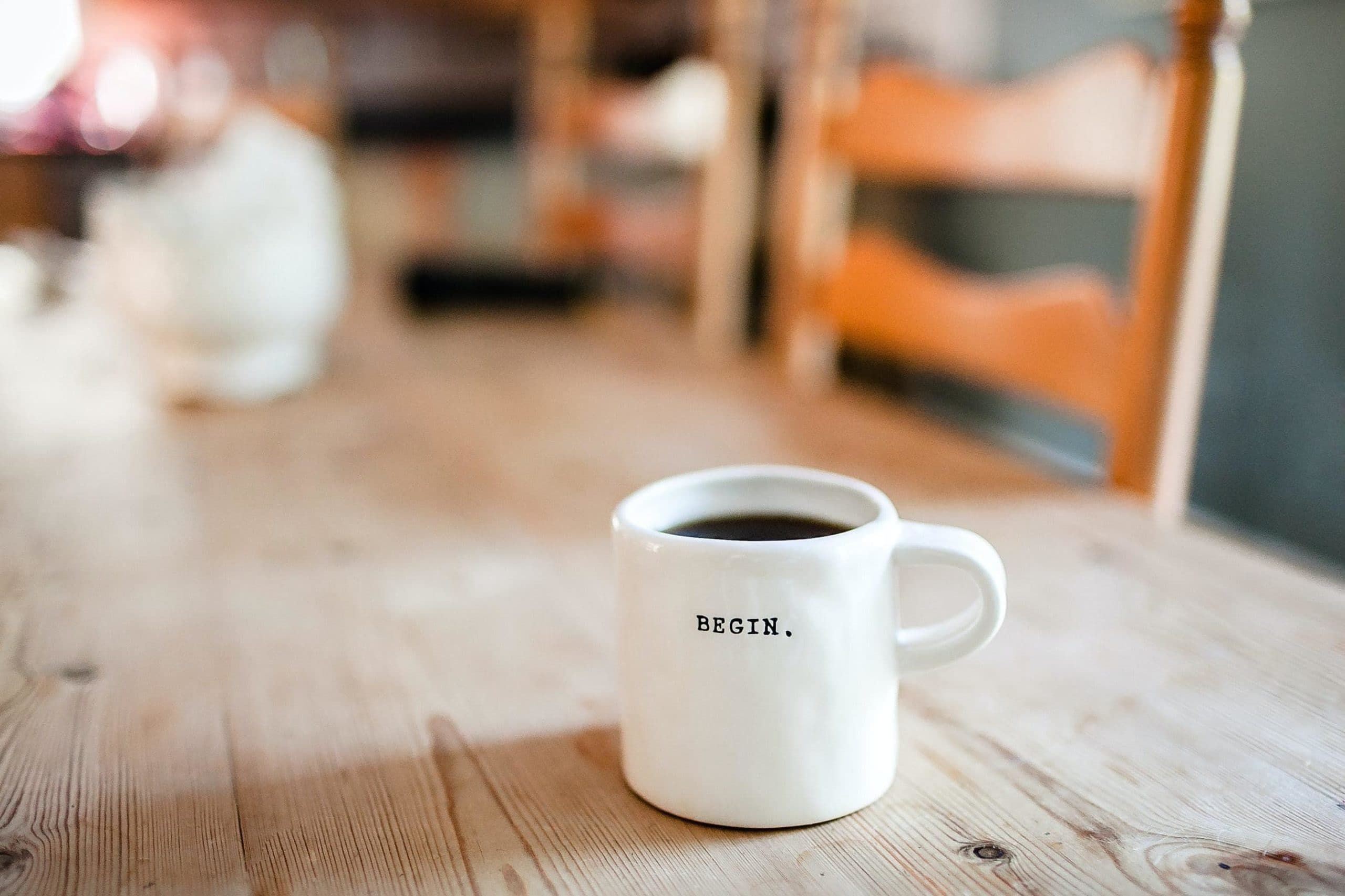 A white mug filled with coffee sits on a wooden table. The mug has the word "BEGIN." printed on it. The background is slightly blurred, focusing attention on the mug.