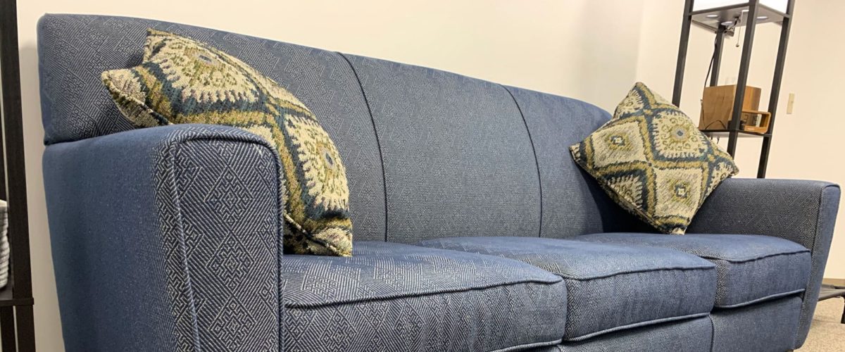 A blue fabric couch with two patterned throw pillows is positioned against a white wall with a metal floor lamp and box in the background.
