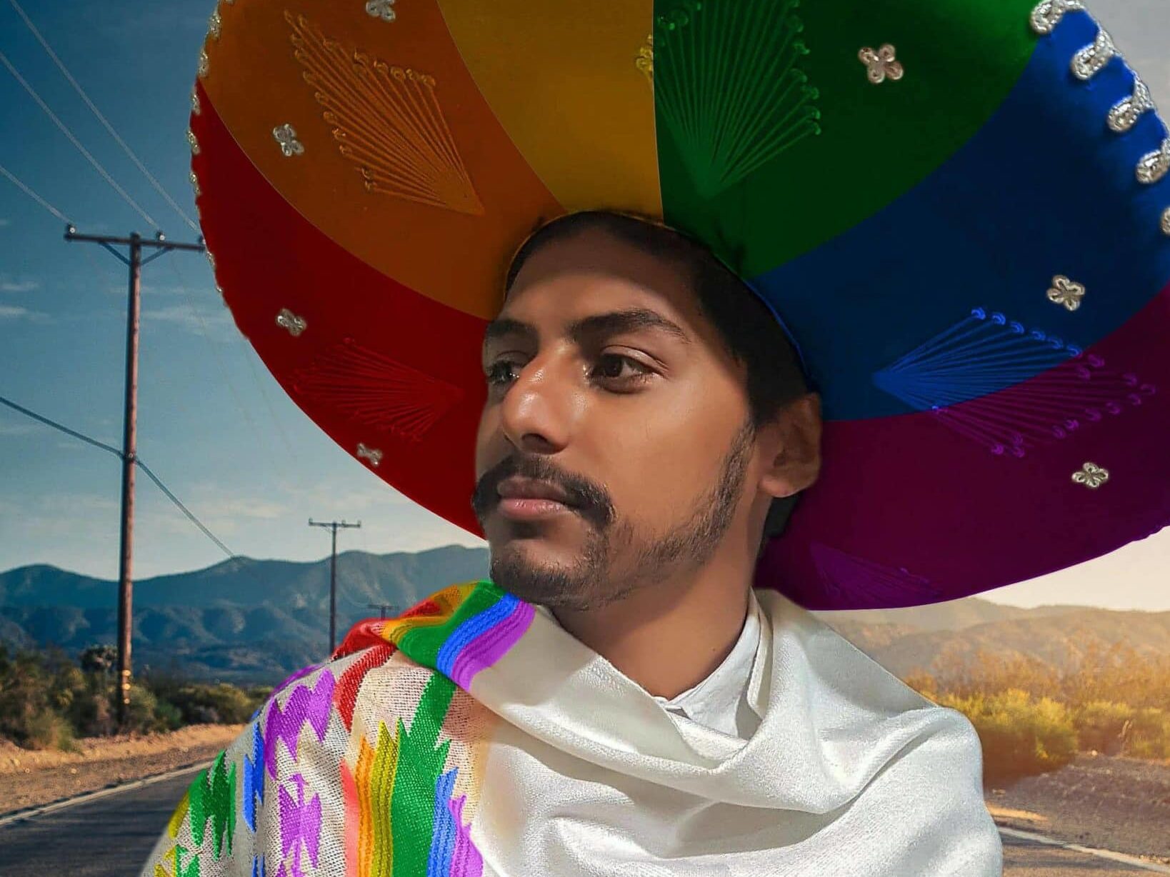 Person with a beard wears a colorful sombrero and a rainbow-patterned garment, looking to the side. Mountains and an open road are in the background.