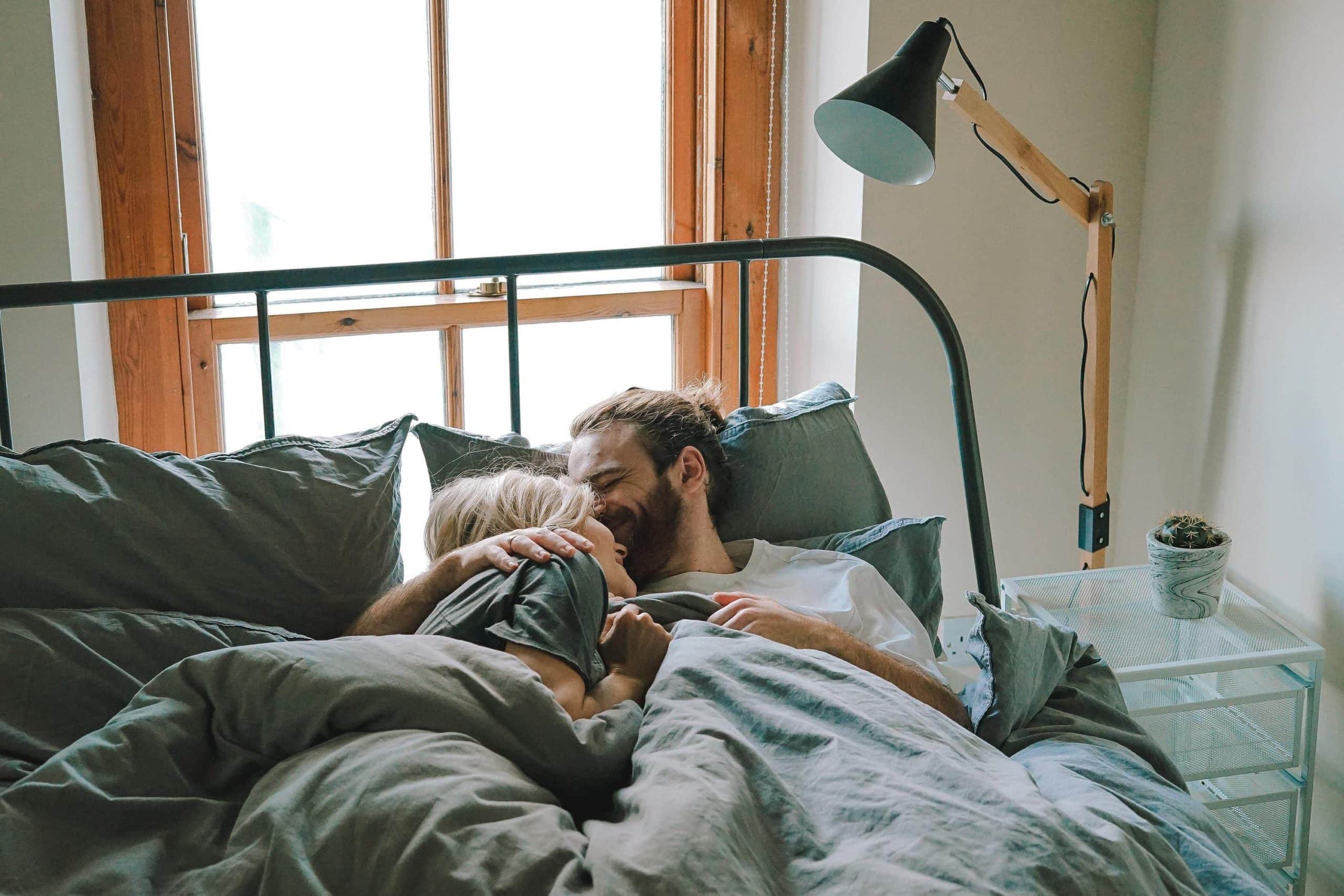 A man and woman lie on a bed, leaning against pillows and laughing. A lamp and potted plant are on a nightstand near the window.