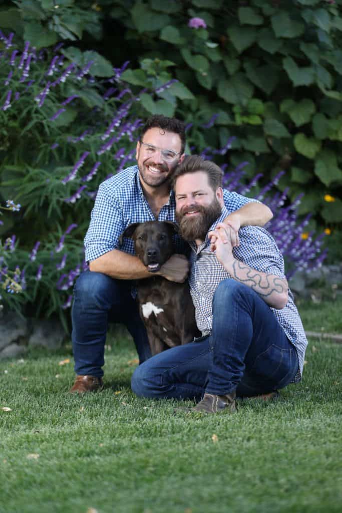 Two men kneel on grass, smiling, with one arm each around a seated black and white dog. They are outdoors with green foliage and purple flowers in the background; it's a picturesque scene likely captured by Kevin Krage.