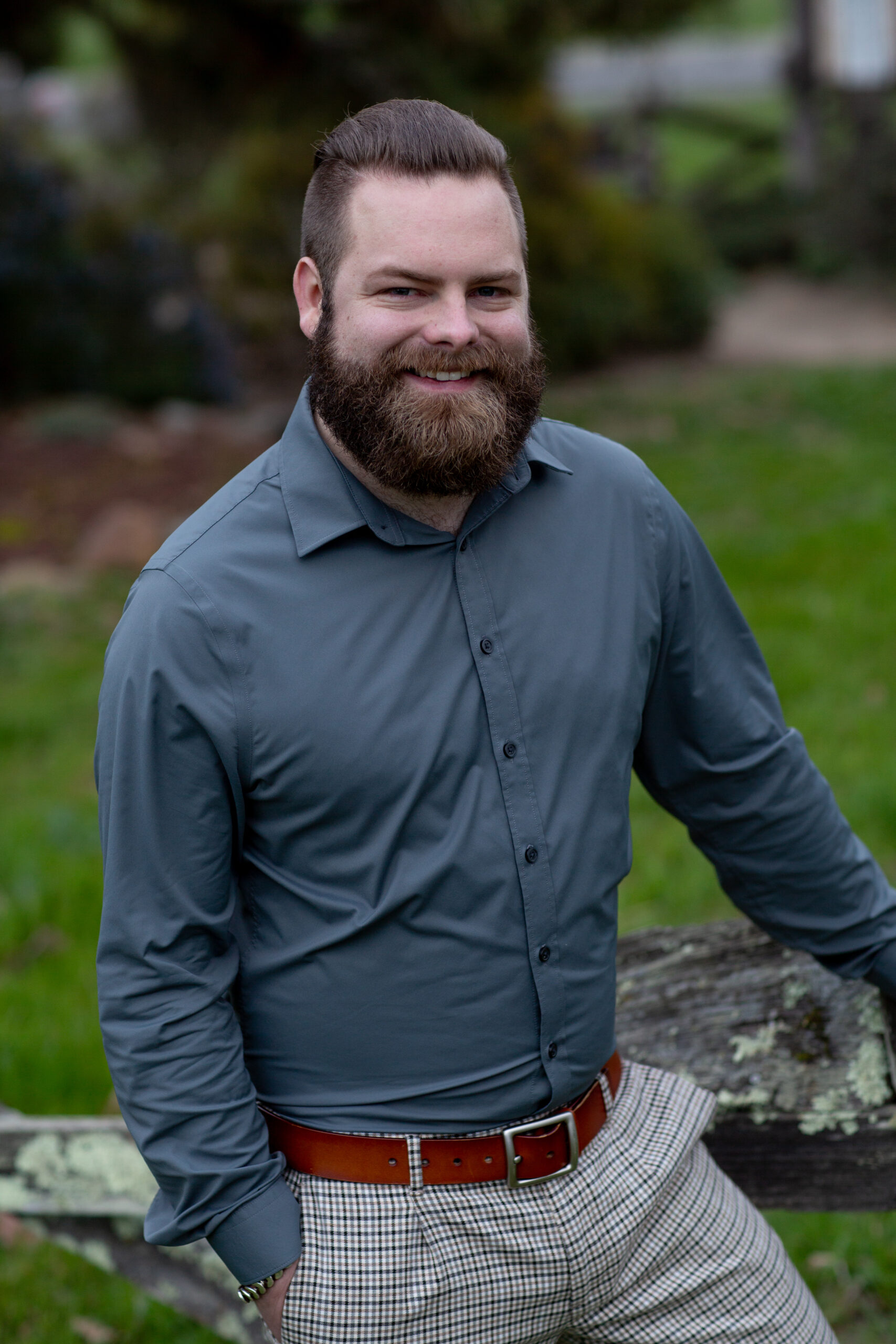 A man with a beard and mustache, wearing a gray dress shirt and checked pants, stands outdoors leaning on a weathered wooden fence, smiling.