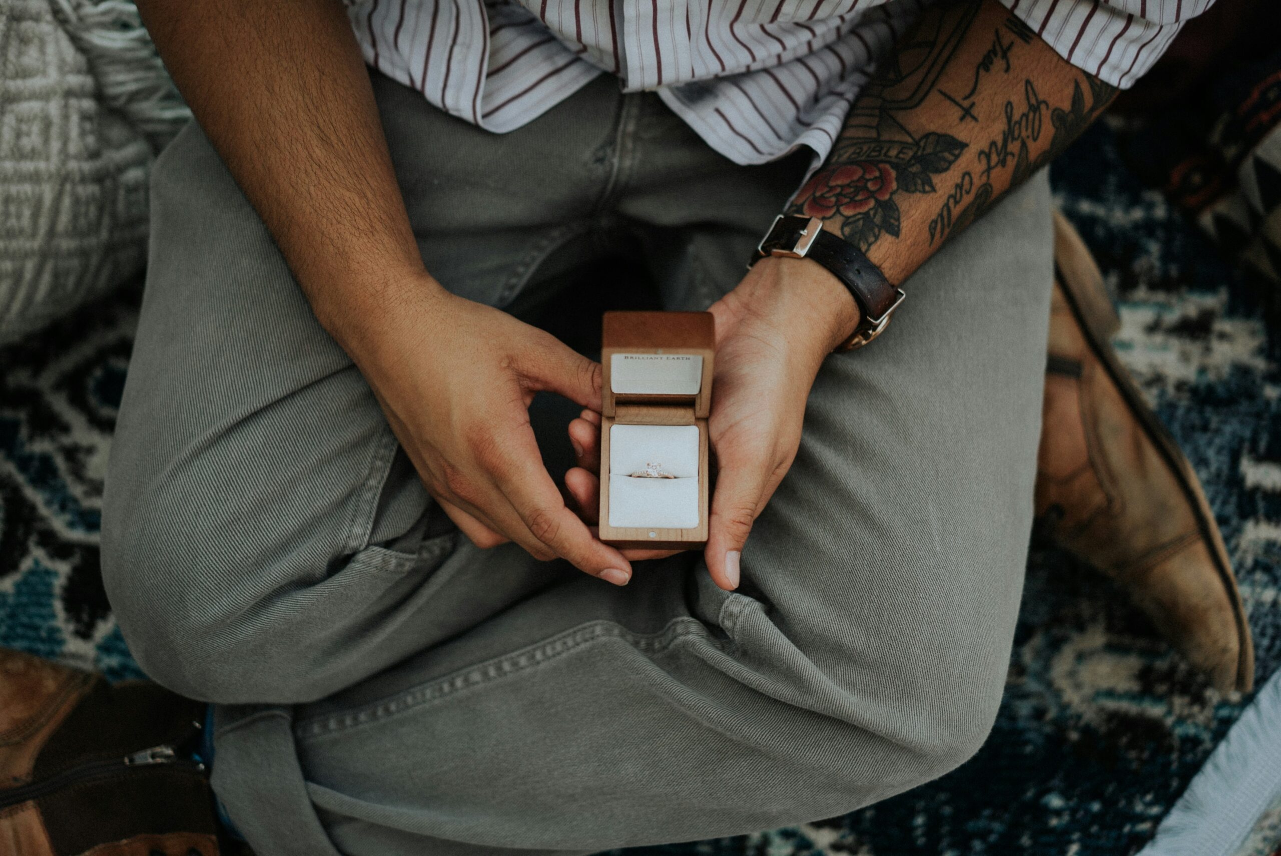 A person wearing a striped shirt and grey pants, with tattoos on their arm, holds an open wooden box containing a ring.