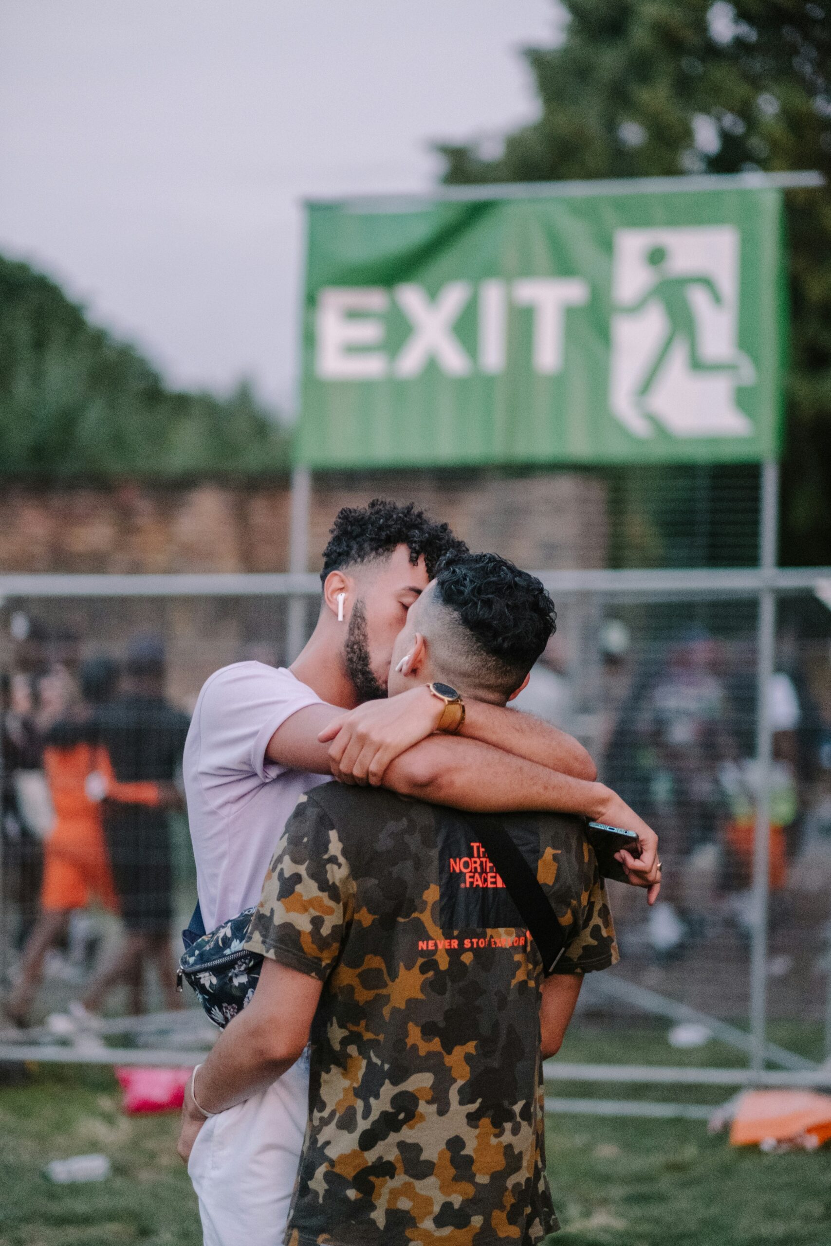 Two people embrace and kiss in front of an exit sign at an outdoor event. One wears a white shirt, and the other wears a camouflage shirt.