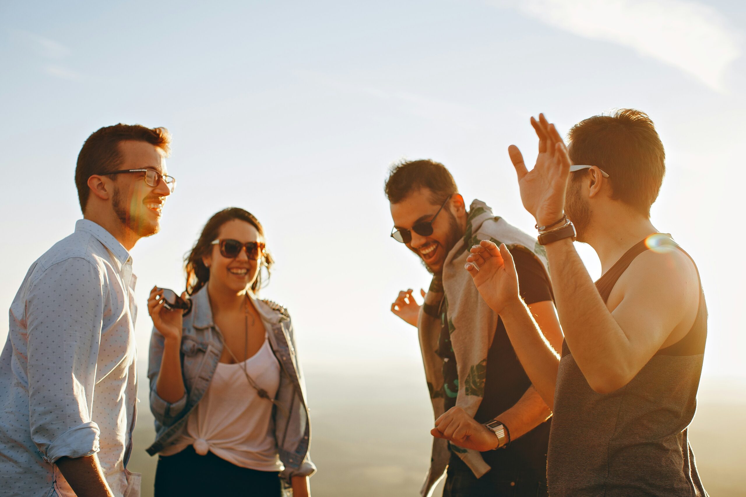 Four people stand together outdoors, laughing and talking, with a bright sky and sunlight in the background.