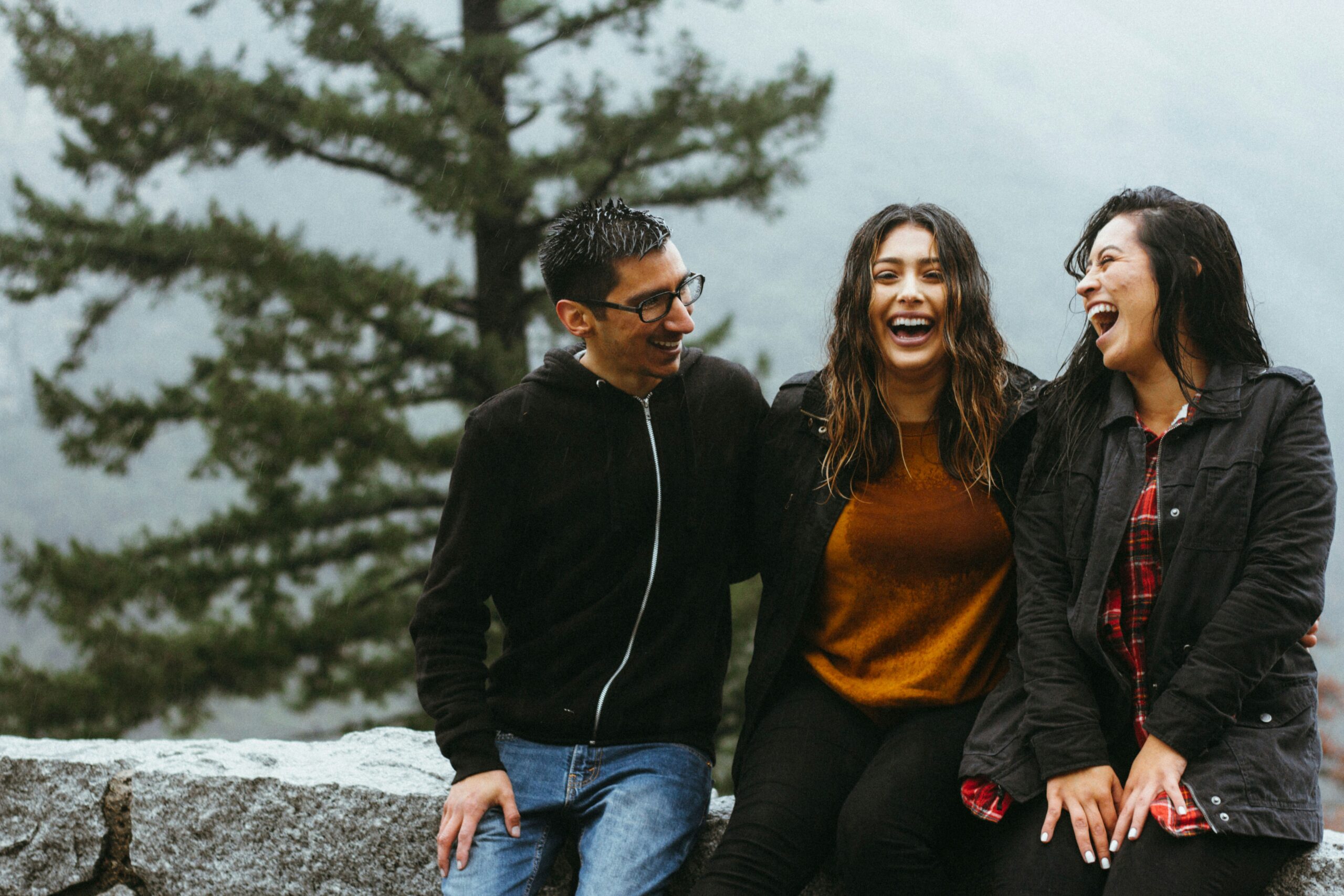 poly relationship with compersion on stone ledge outdoors, smiling and laughing together. Trees and a foggy background can be seen behind them.