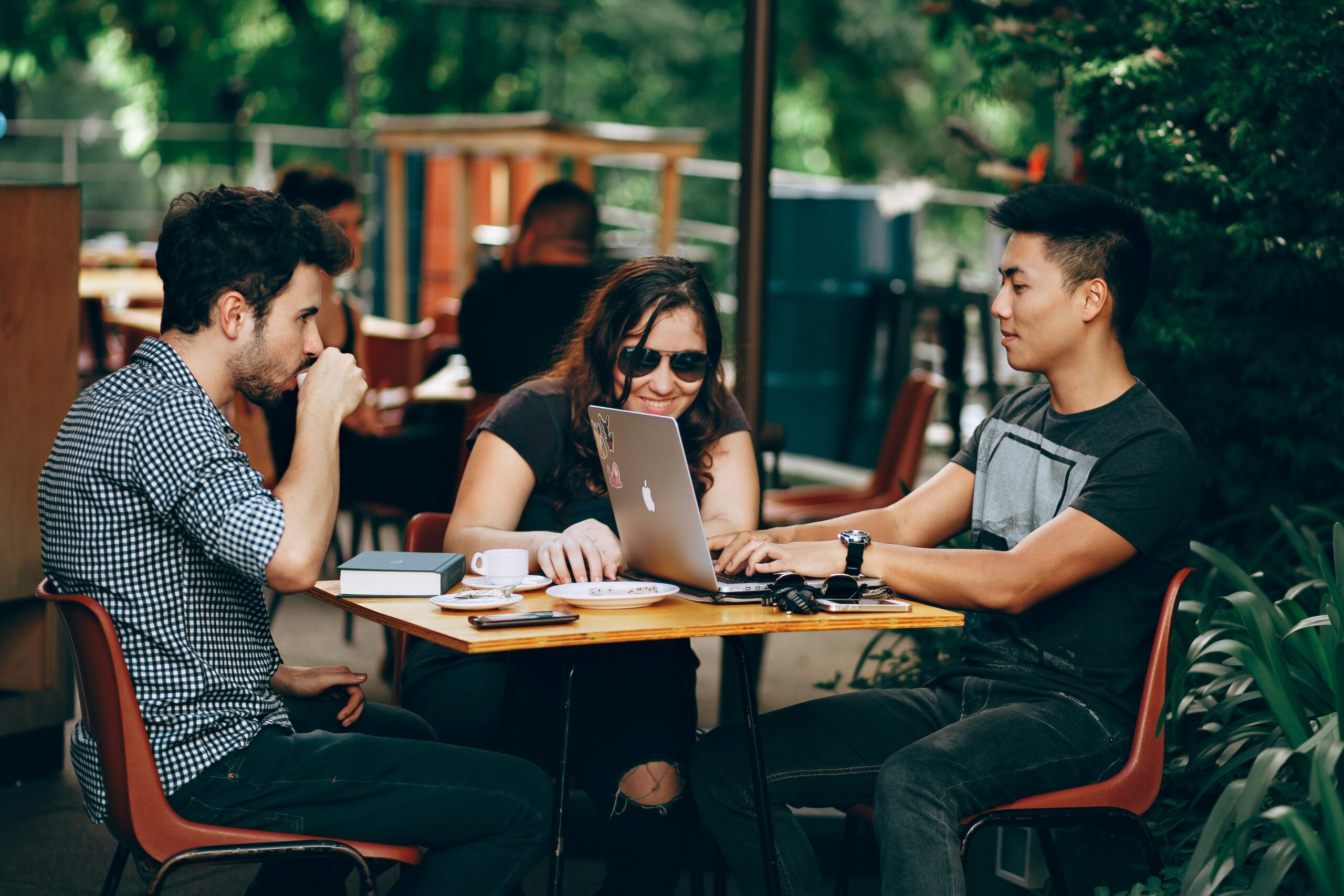 Three people sit at an outdoor café table, engaged with a laptop. Two men face each other, with a woman between them, smiling. Coffee cups and books are on the table.