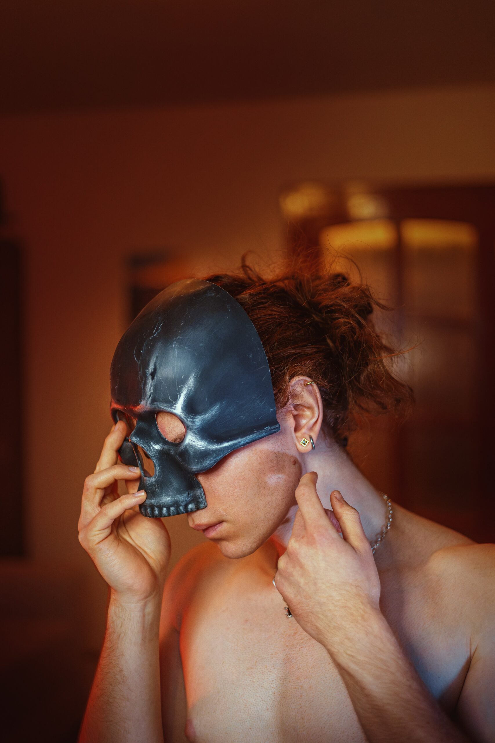 Person wearing a black skull mask, adjusting it with one hand. Shirtless, with curly hair tied back and an earring visible. Dimly lit background.