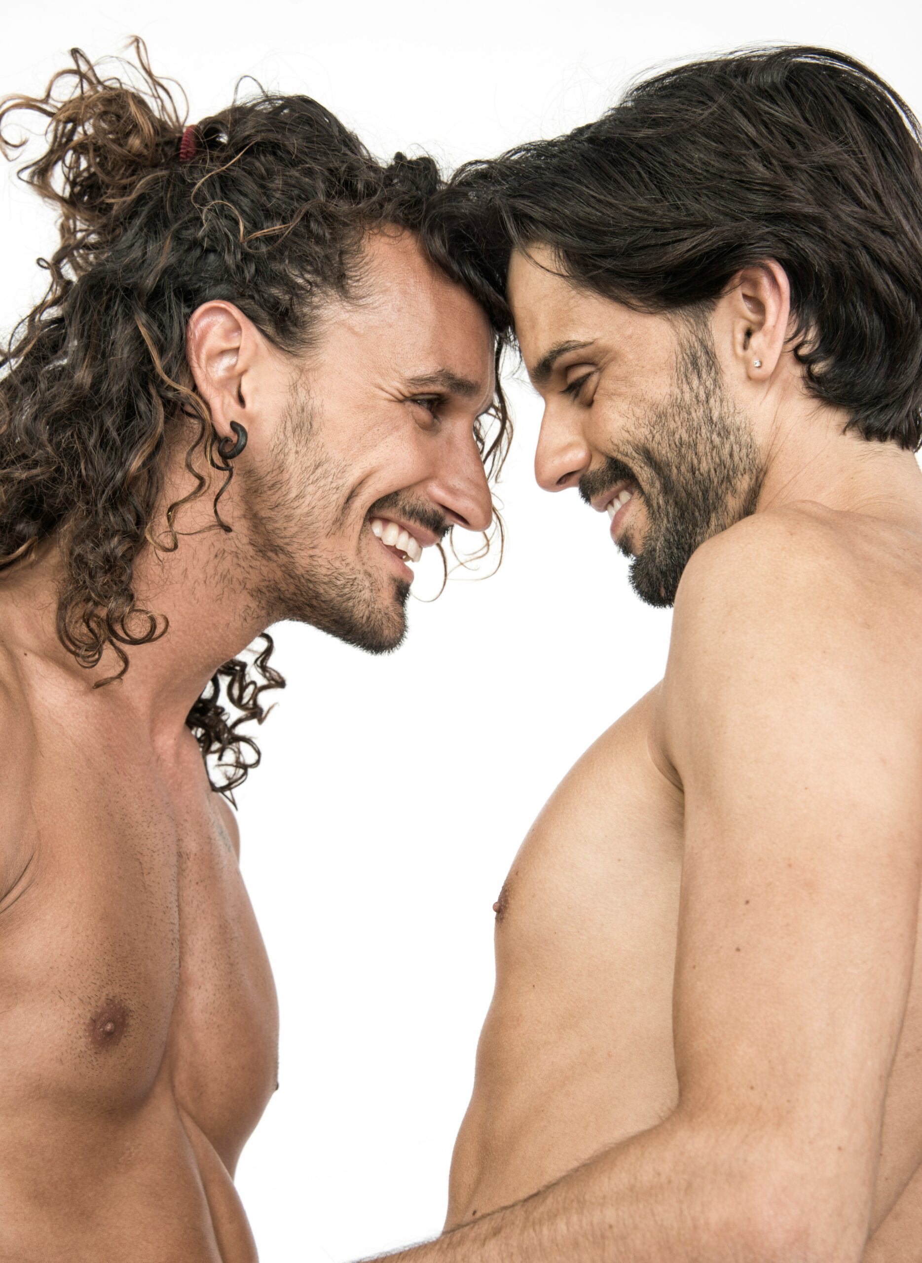 Two shirtless men with long hair face each other, smiling, against a white background.