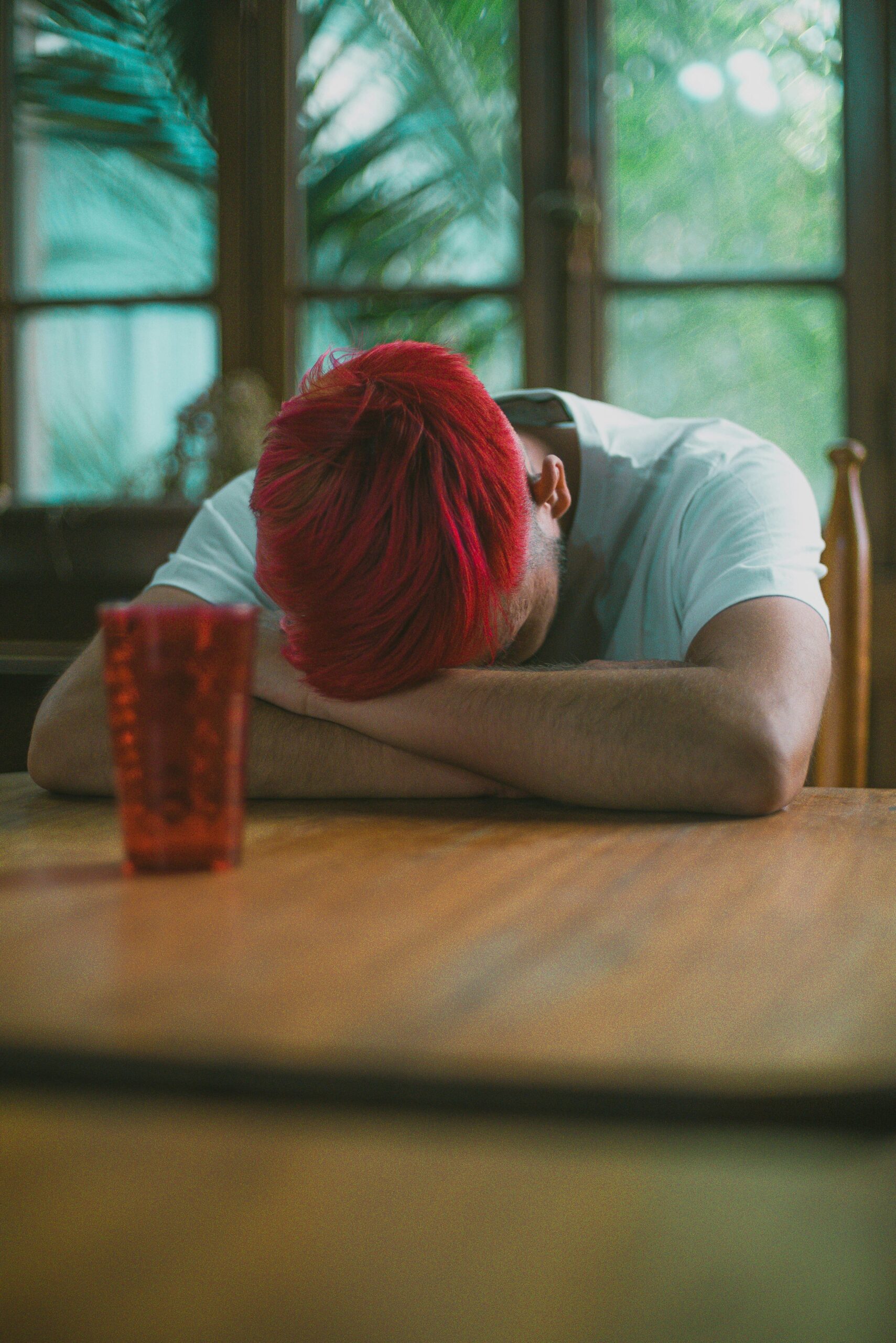 Person with red hair rests their head on crossed arms at a wooden table in a room with green plants outside the window and a red glass nearby.