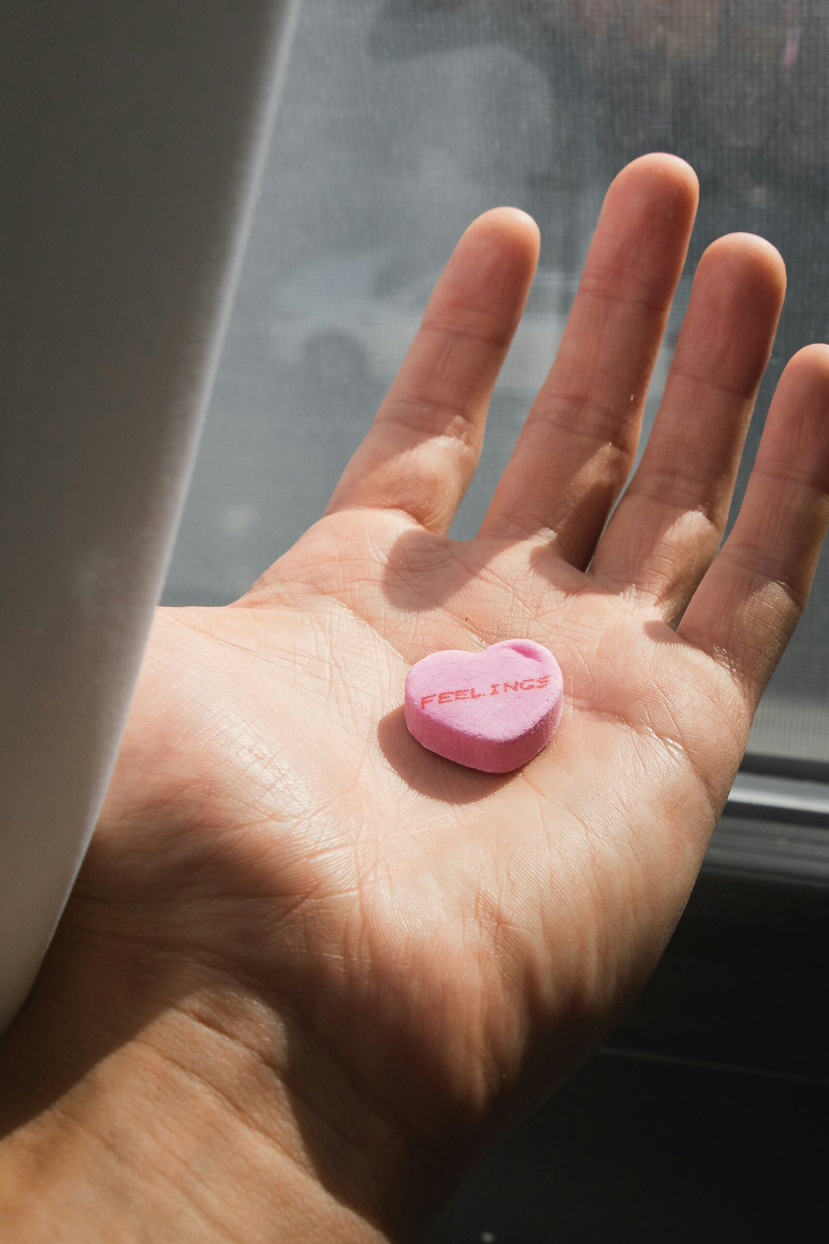 A hand holds a pink candy heart with the word "FEELINGS" in a sunlit setting by a window.