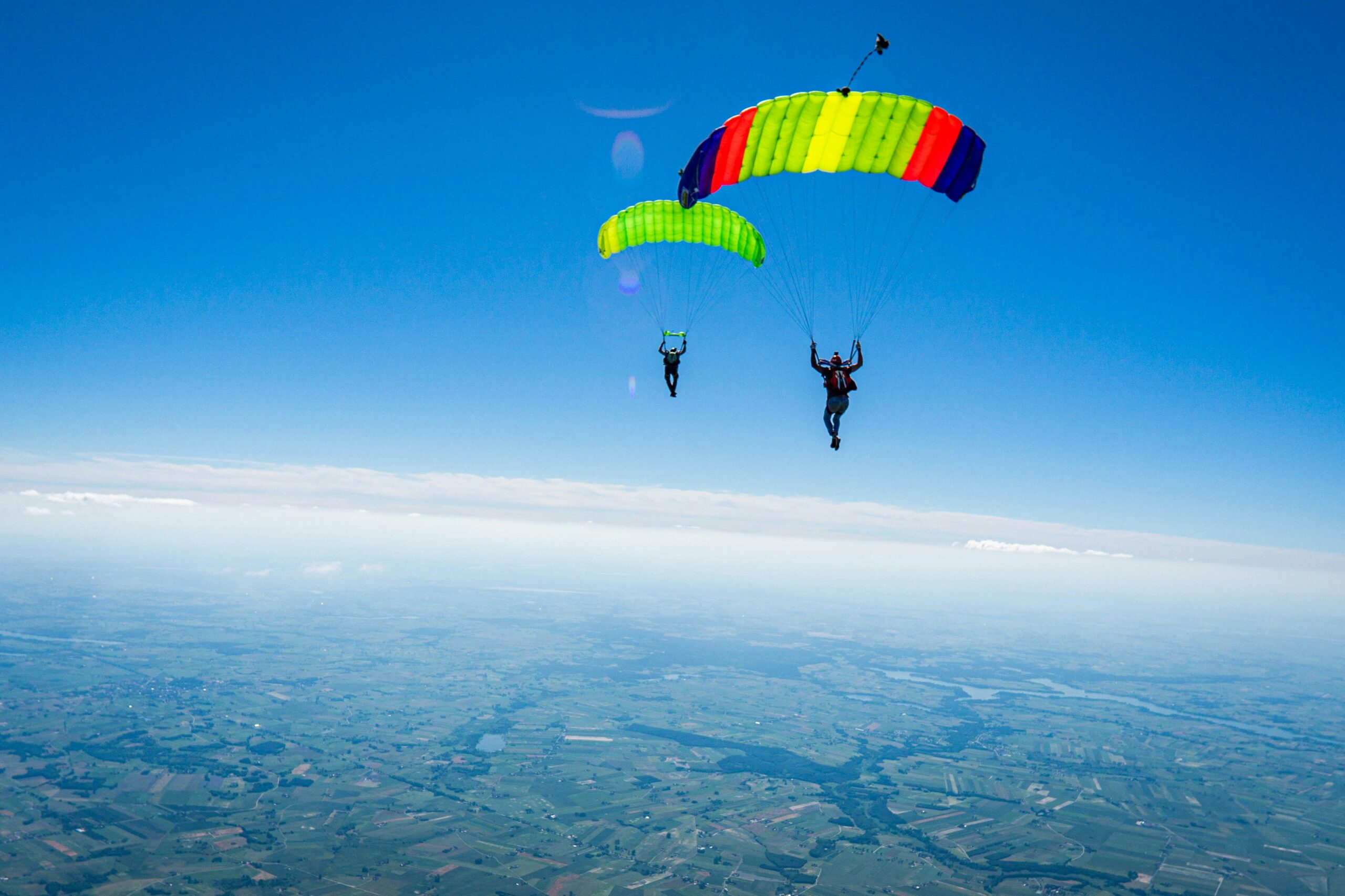 Two skydivers with colorful parachutes descend against a clear blue sky over a vast landscape.