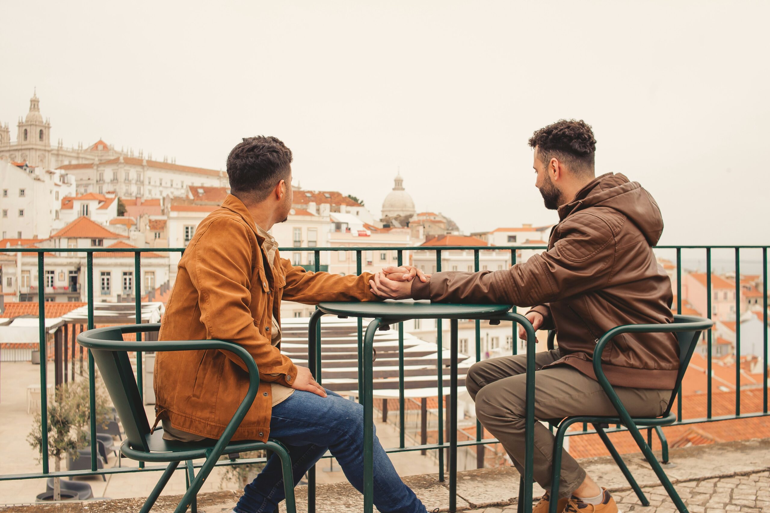 Two people sit at an outdoor table holding hands, overlooking a cityscape with traditional buildings and a distant dome.