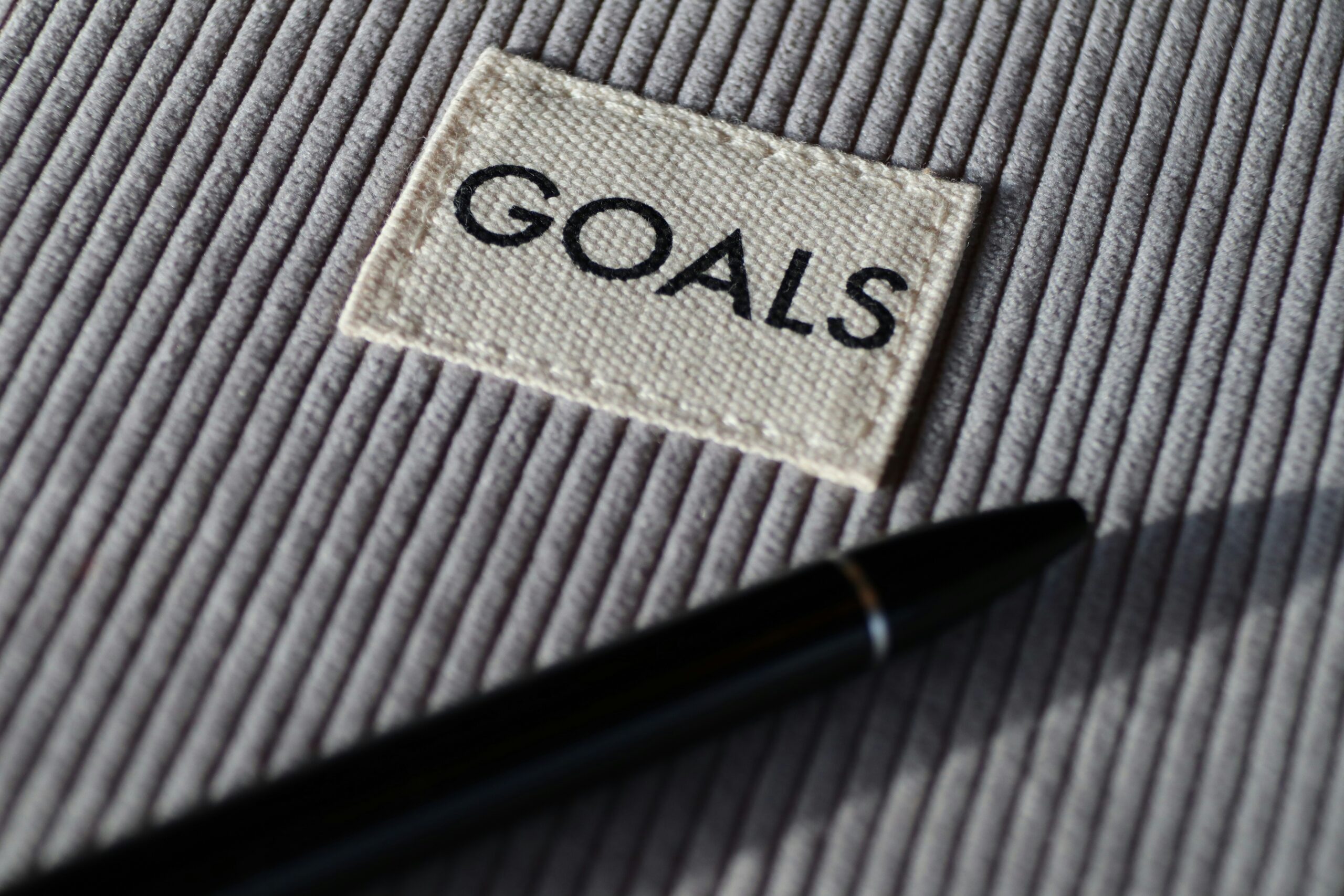 A pen rests on a textured surface next to a fabric patch labeled "GOALS.