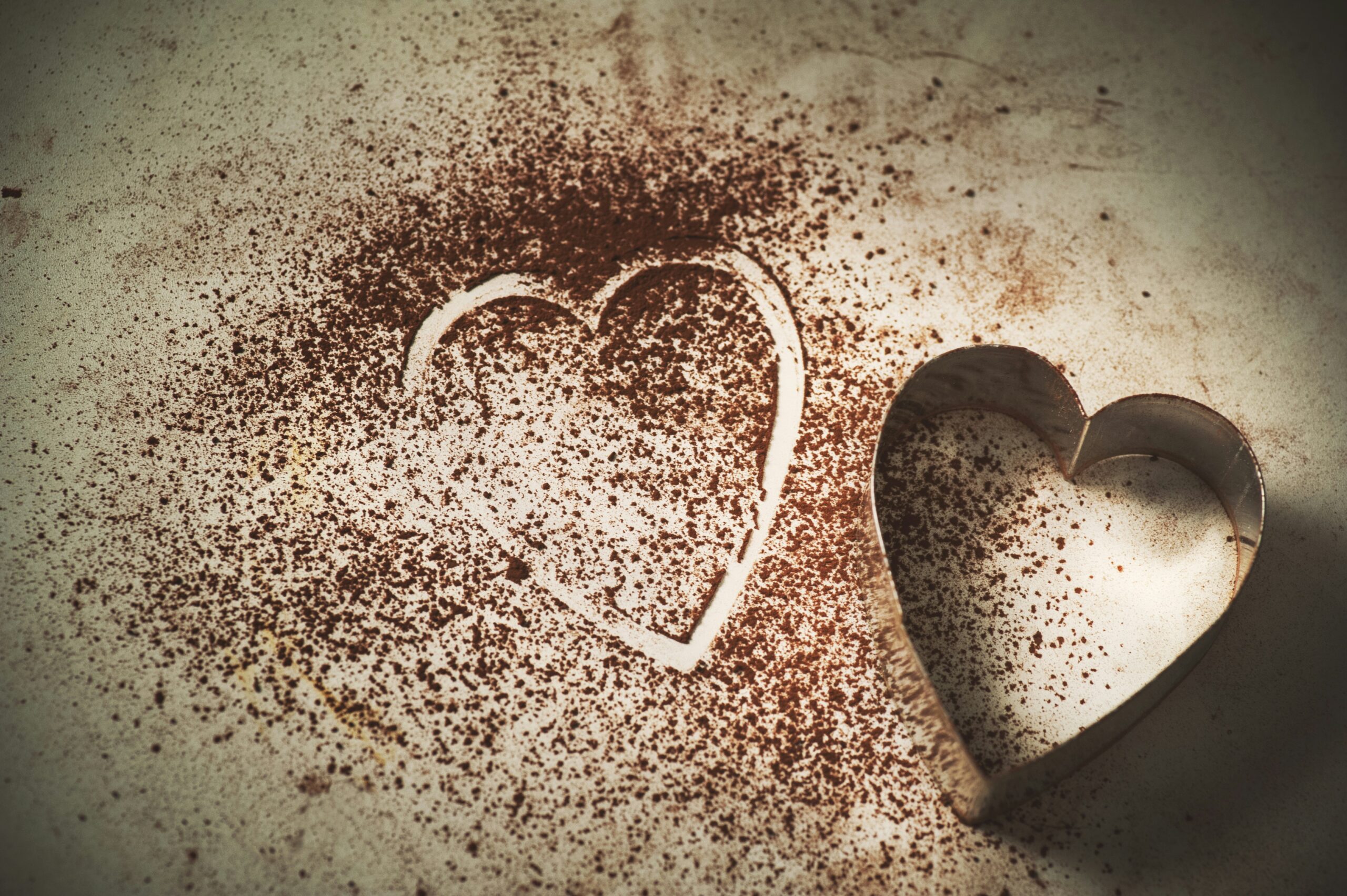 A heart-shaped cookie cutter on a surface with cocoa powder, leaving an outline of a heart next to it.