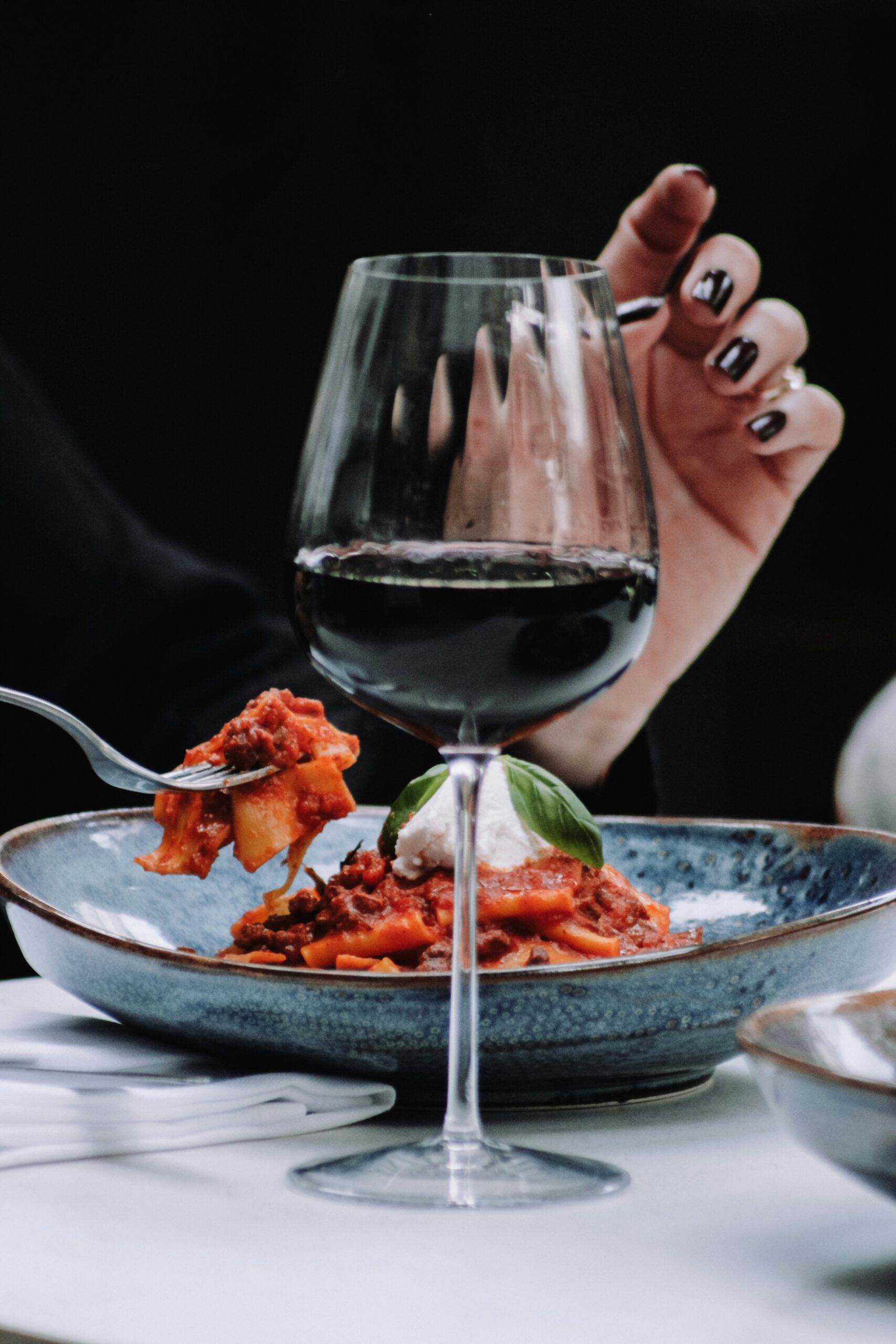 A person's hand holding a fork with pasta, in front of a glass of red wine on a table.