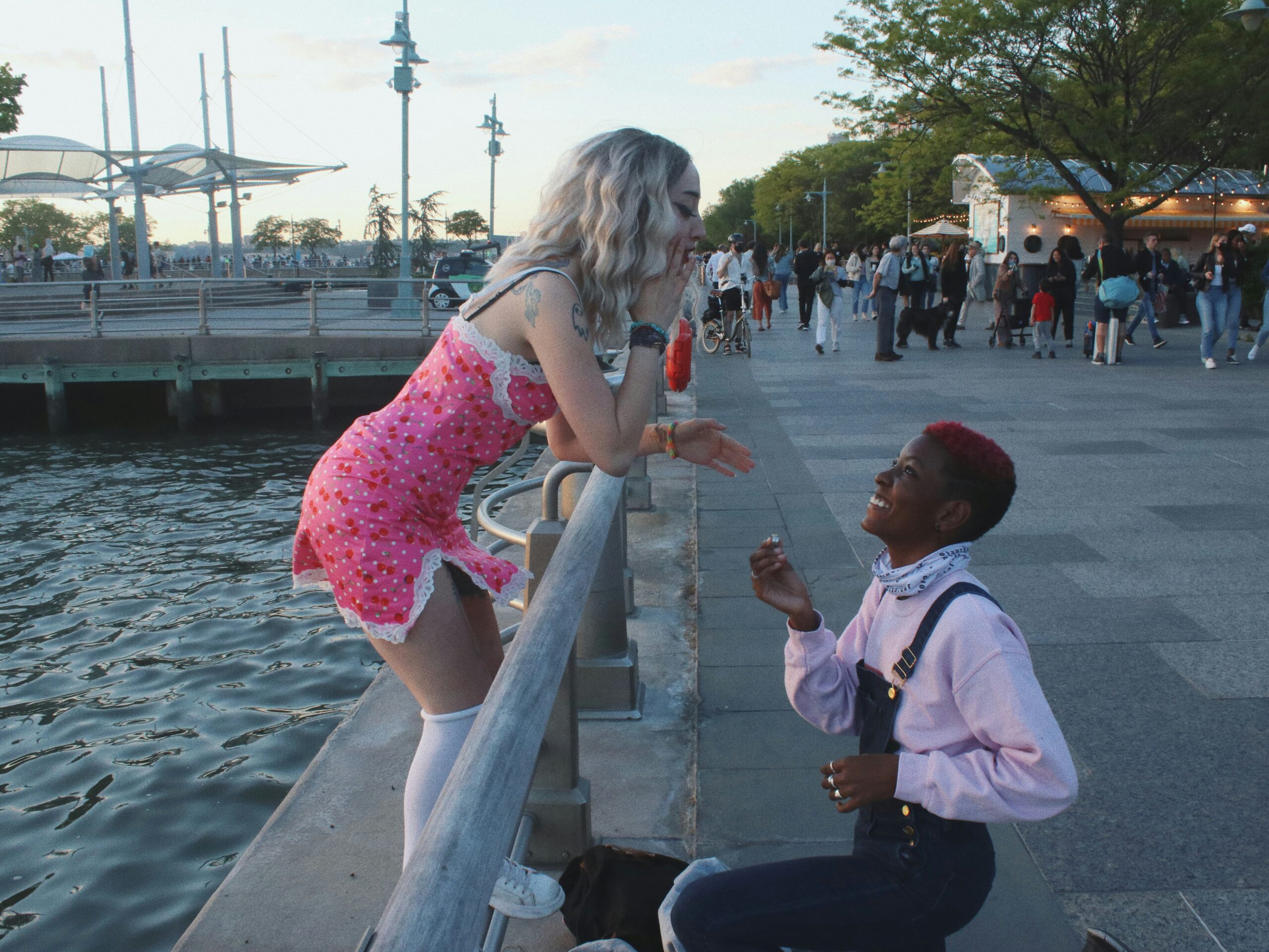Two people are at a waterfront promenade. One person is kneeling, holding a ring box, while the other is surprised and laughing. Onlookers in the background.
