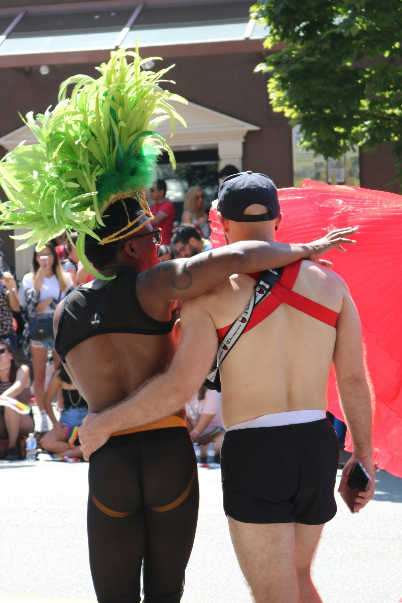 Two people in colorful outfits, one with a large green headdress and the other with a red cape, walk arm in arm during a parade. People are watching in the background.