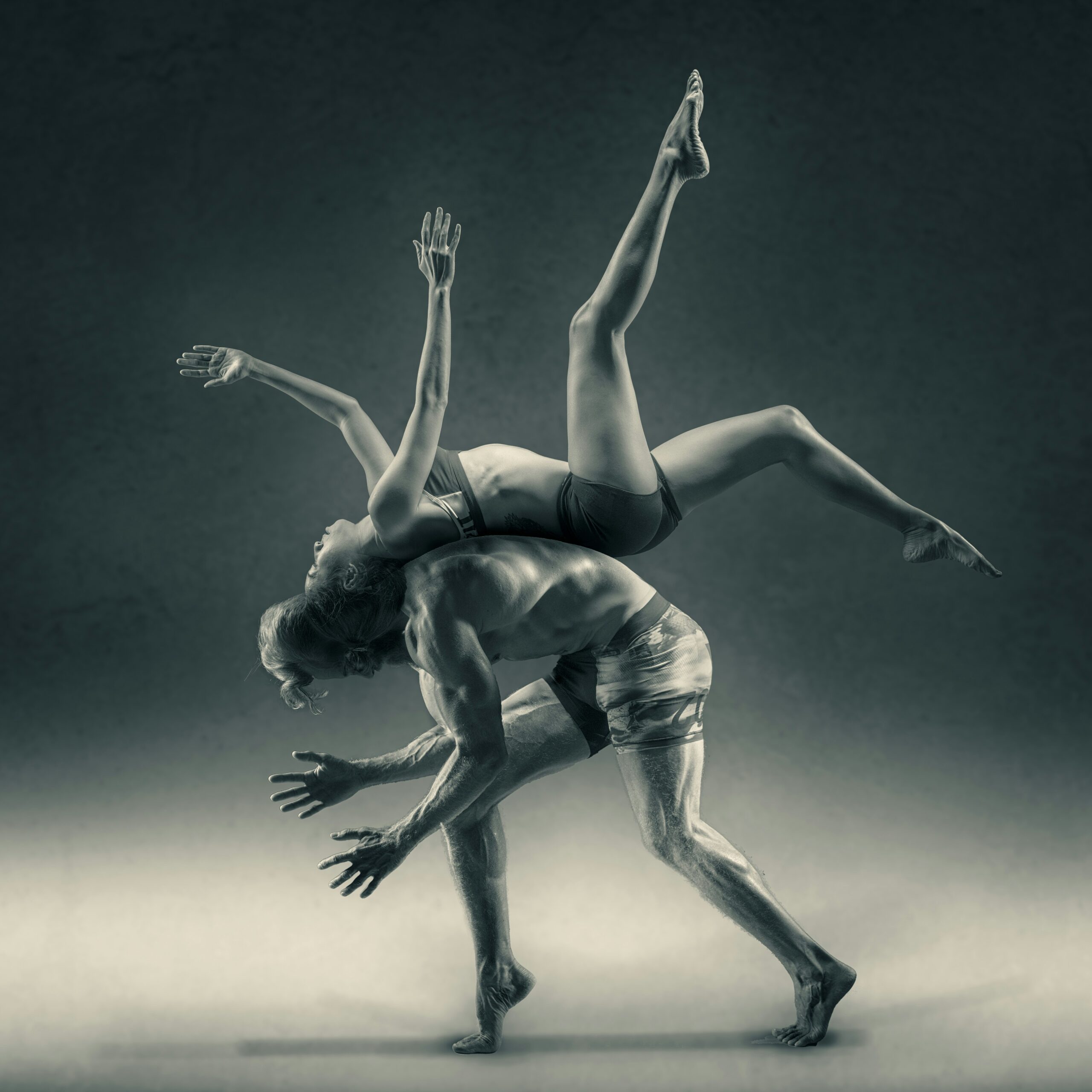 Two dancers perform an acrobatic pose with one balanced on the back of the other, both with arms outstretched.
