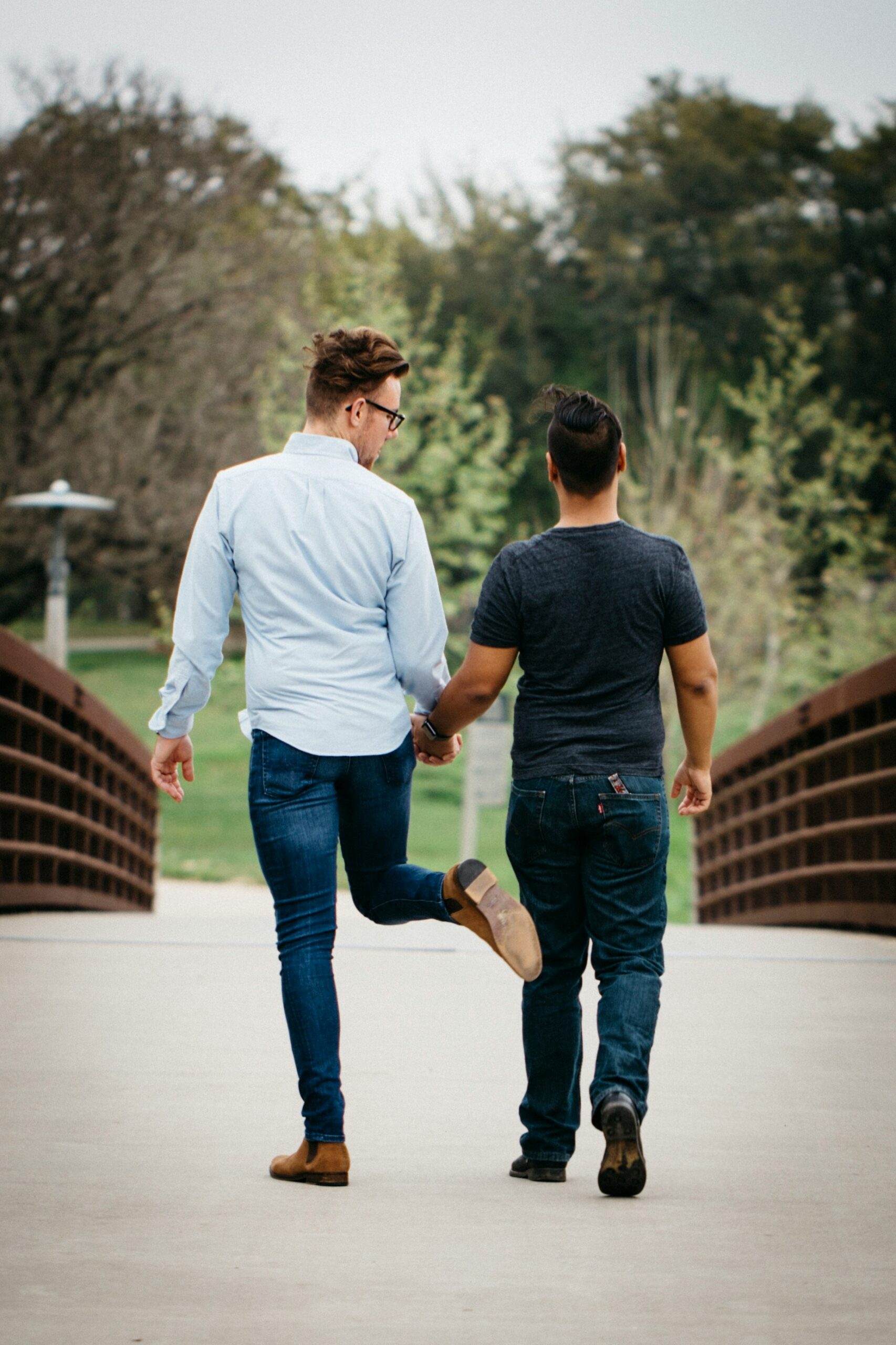 Two people holding hands walk away on a bridge. One lifts a leg playfully. Trees and greenery are in the background.