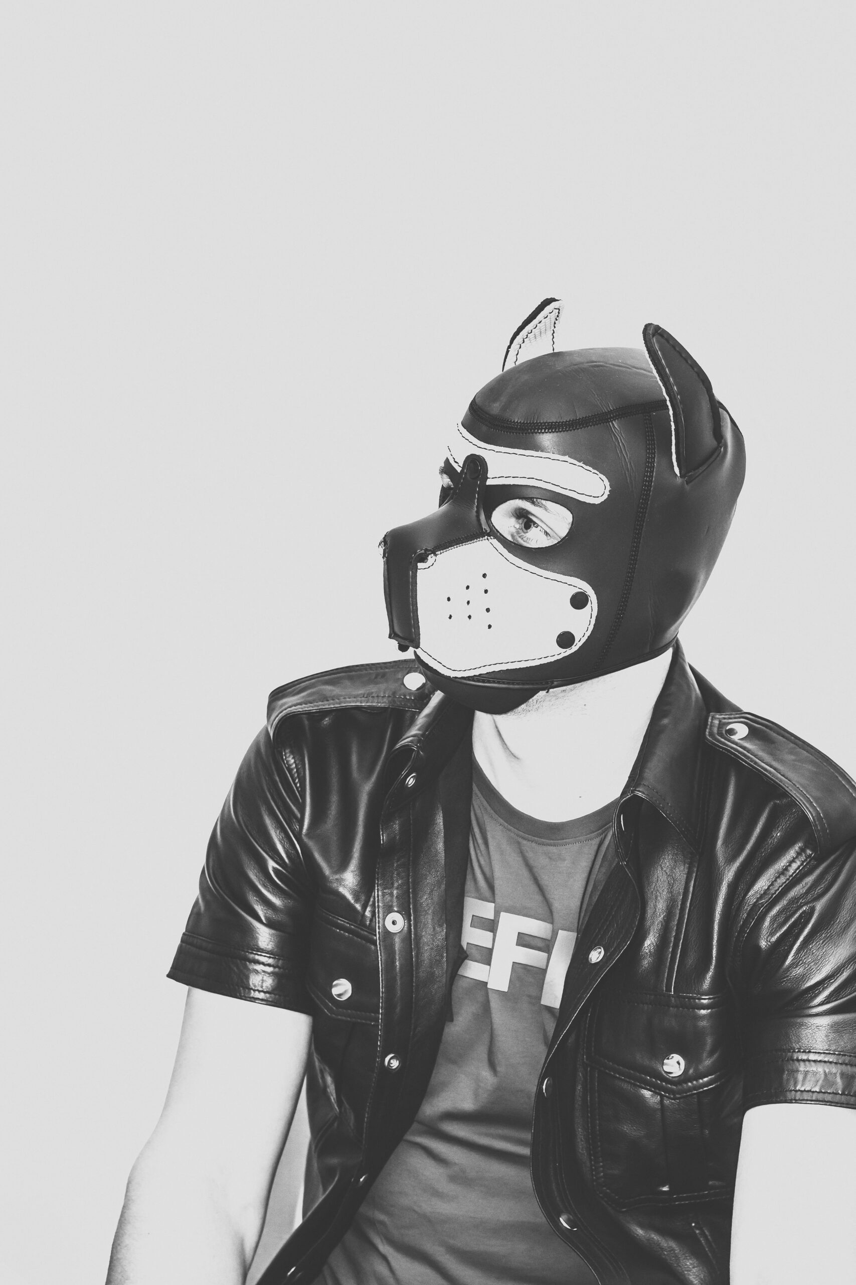 Person wearing a black leather dog mask and leather shirt, sitting against a plain background.