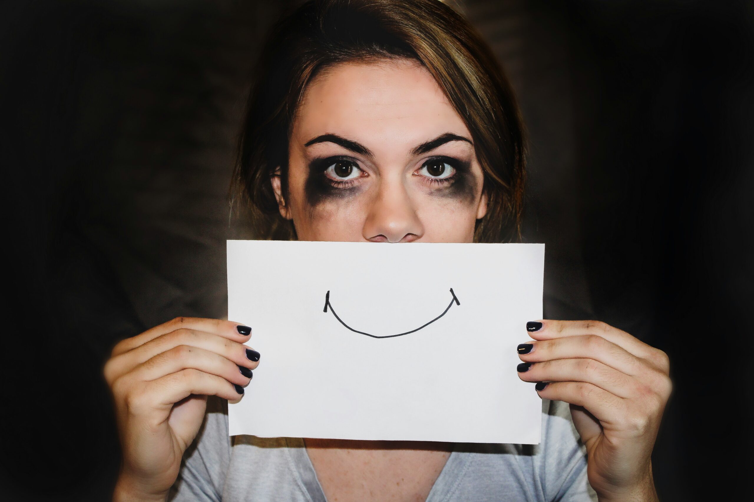A person with smudged eye makeup holds a paper with a drawn smile in front of their mouth.