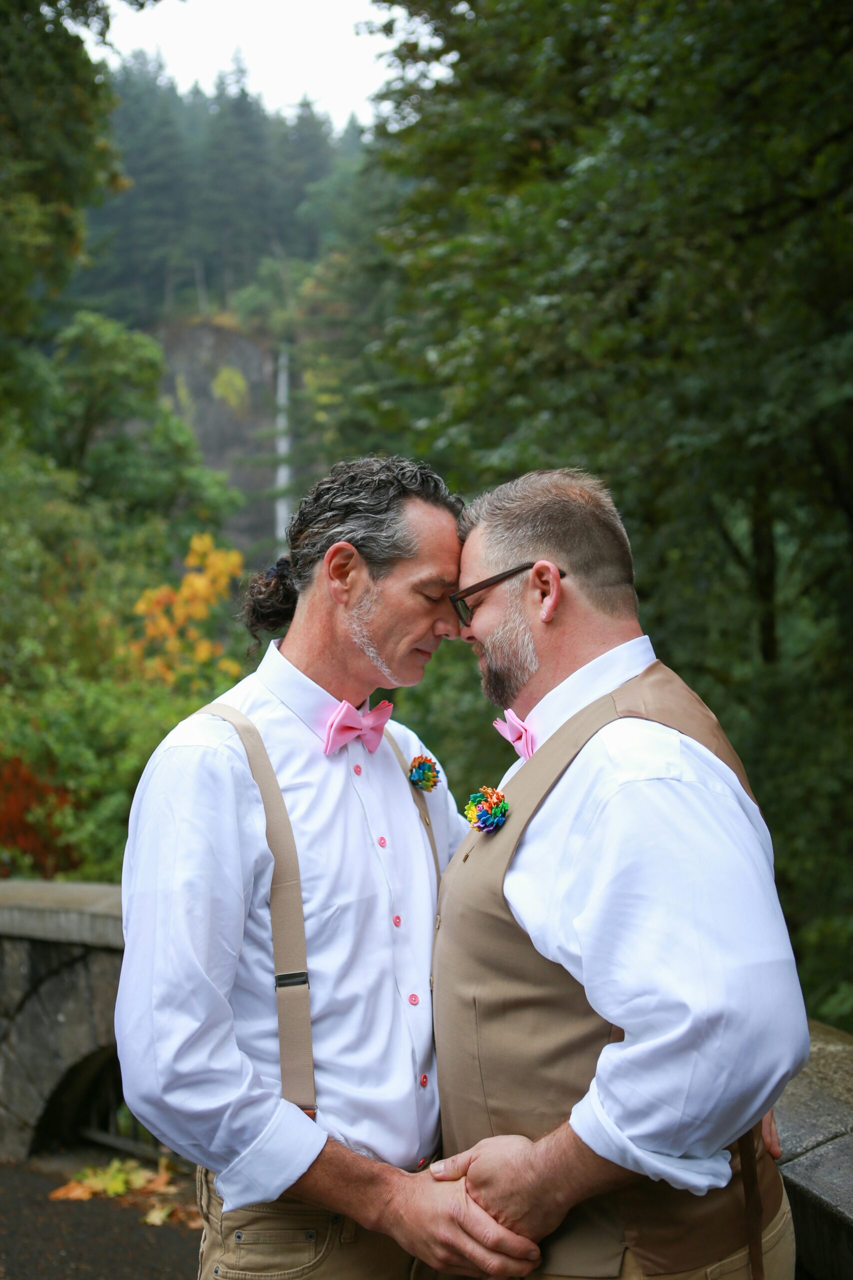 Two men dressed in white shirts, tan vests, and pink bow ties stand close, touching foreheads, holding hands outdoors with greenery in the background.